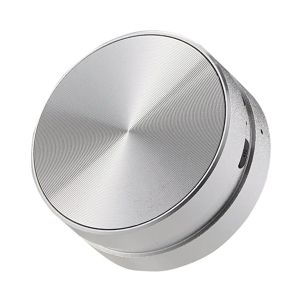 A compact and sleek SoundSurface Mini Resonance Speaker made of aluminum, displayed on a wooden surface, with a plant and a notebook in the background.