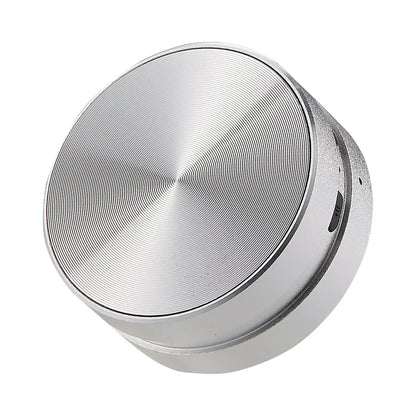 A compact and sleek SoundSurface Mini Resonance Speaker made of aluminum, displayed on a wooden surface, with a plant and a notebook in the background.